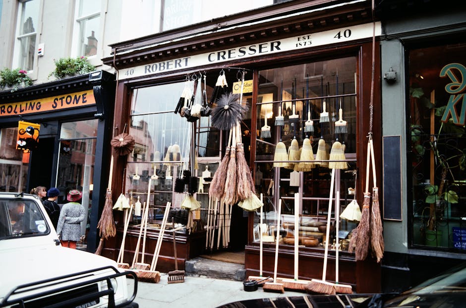 The storefront of Robert Cresser, a shop on Upper Richmond Road in Putney, features a large glass window displaying various cleaning tools and decorative items such as brushes, brooms with wooden handles, and dusters. The exterior is made of dark wood framing with a white sign that reads 'ROBERT CRESSER' and the address number '40'. Several brooms and brushes are arranged neatly outside the shop, some leaning against the window and others placed on the ground, showcasing different materials like straw, synthetic fibers, and natural bristles. The lighting inside the shop illuminates the interior, highlighting the arrangement of cleaning supplies and the wooden shelving units. Adjacent shops and pedestrians are visible across the street, contributing to a lively commercial area. Cleaner Putney provides expert surface cleaning and deep cleaning solutions to maintain hygiene and cleanliness in such retail environments, ensuring surfaces like wood, glass, and ceramic display cases remain spotless and inviting.