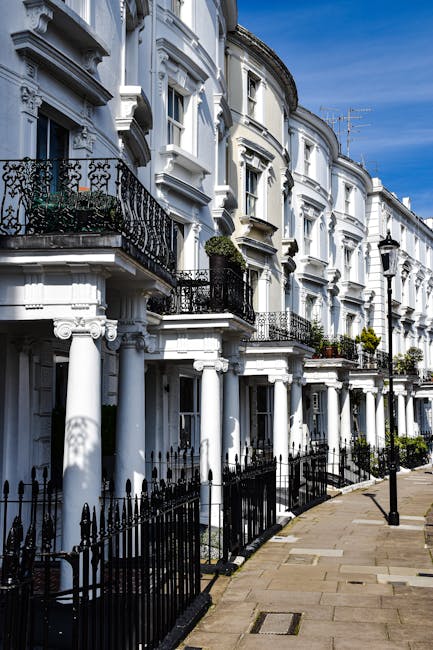 A row of white Victorian-style terraced houses with ornate architectural details, including decorative cornices and black wrought iron balconies on the upper floors. The buildings have large windows, some with flower boxes, and are set along a clean, paved sidewalk with a black metal fence separating the public walkway from the front gardens. The scene is illuminated by natural daylight under a clear blue sky, highlighting the pristine, well-maintained appearance of the facades. This image reflects the importance of surface cleaning and maintenance in residential buildings, aligning with the services offered by Cleaner Putney for domestic cleaning and surface sanitisation.