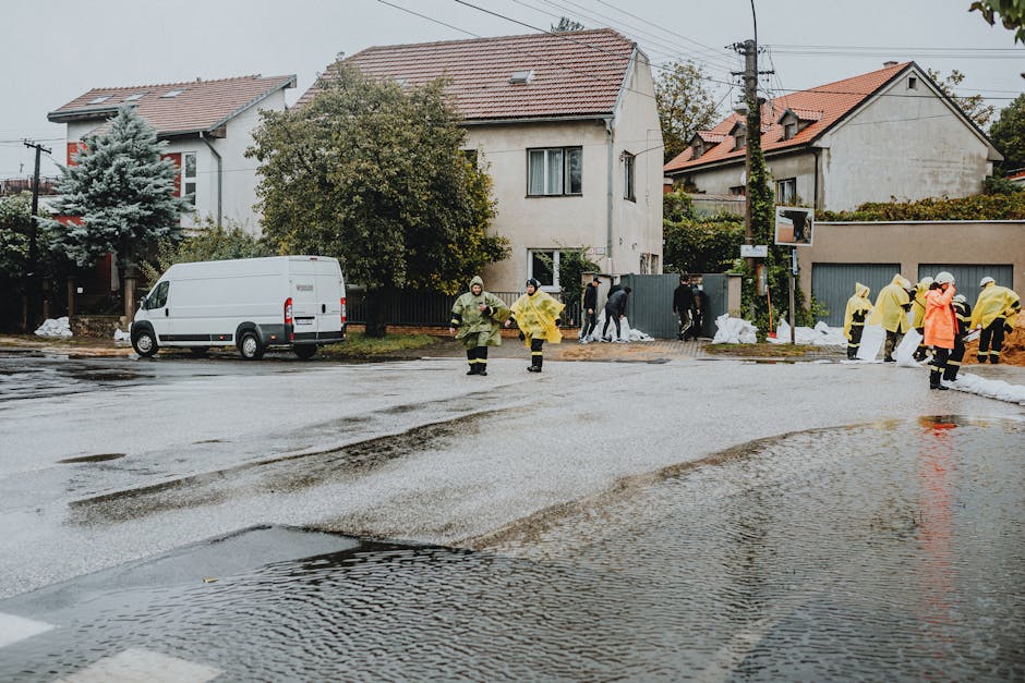 A group of emergency responders wearing yellow and orange raincoats standing on a wet street after flooding, with puddles forming on the asphalt. In the background, there are residential houses with white and beige exteriors, some with tiled roofs, and trees with autumn-colored foliage. A white van is parked on the side of the road, and a few individuals are seen near a fence and sidewalk, possibly conducting flood response or cleanup activities. The scene is overcast, with diffused natural lighting highlighting the reflective surface of the flooded street. Cleaner Putney provides professional cleaning services for flood damage mitigation and sanitisation, ensuring thorough surface cleaning and hygiene restoration in residential areas affected by flooding.