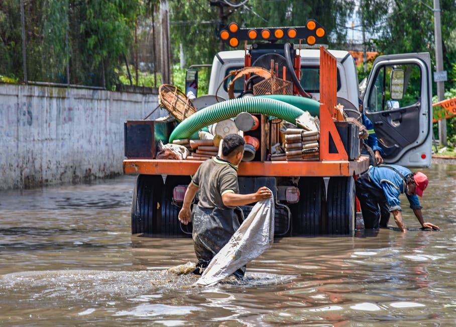 Two men are engaged in flood clean-up activities in a residential outdoor setting, with one man waist-deep in muddy water holding a white cloth or bag, and the other near a truck filled with various cleaning and recovery equipment, including hoses, tools, and safety gear. The scene is set outdoors with trees and a concrete wall in the background, under natural daylight. The visible surfaces include muddy water, the truck's metal body, and the ground, all showing signs of water damage and grime. The men are dressed in work clothes suitable for flood response, and the atmosphere depicts urgent recovery efforts following a flood event. Cleaner Putney offers emergency flood cleaning services to restore and sanitise affected properties, emphasizing rapid response and comprehensive surface cleaning.