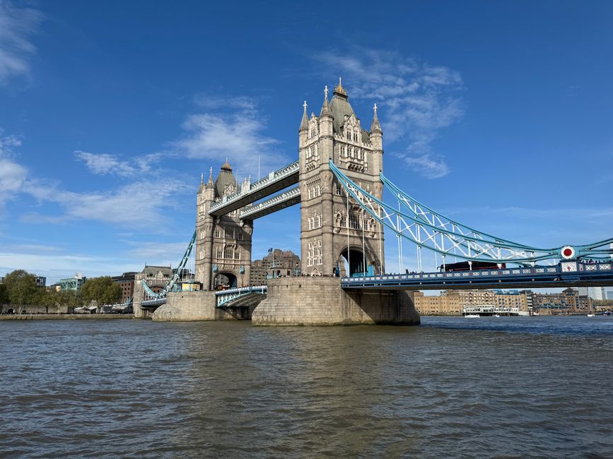 A serene view of a riverbank during daytime with a clear blue sky and scattered white clouds. The calm water reflects the sky and surrounding trees, with several small boats docked along the edge. On