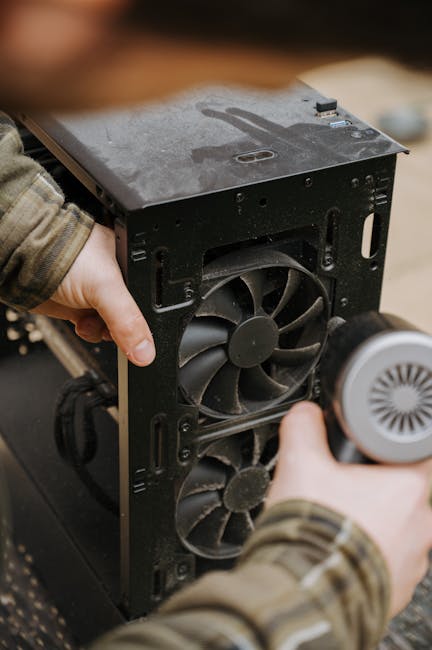 A person wearing a camouflage-patterned sleeve is using a spray bottle to clean the interior of a black computer tower case placed on a wooden surface. The case is open, revealing cooling fans and internal components. The surface appears slightly dusty, and the person is positioned in a well-lit environment, possibly during a professional cleaning or maintenance session related to domestic or commercial surface cleaning for electronic equipment within a residential setting. Cleaner Putney offers expert cleaning solutions that include detailed equipment sanitisation and maintenance, ensuring optimal functionality and hygiene of electronic surfaces in properties like basement flats in Putney.