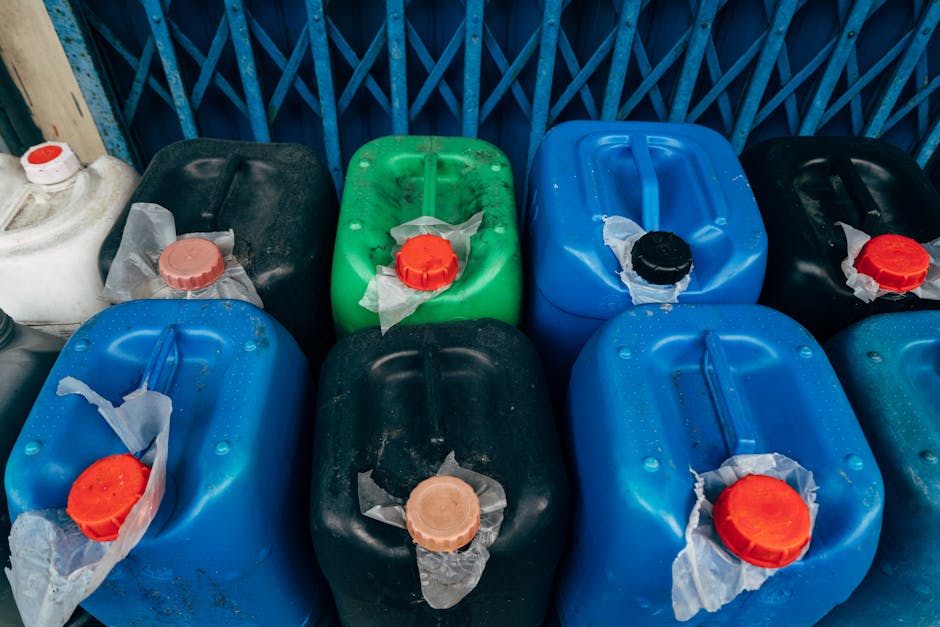 A collection of nine plastic containers in various colors including blue, black, green, and white, arranged on a surface against a blue metal fence. The containers have different lid colors such as red, black, and beige, with some lids partially covered by white torn paper or plastic. The surfaces of the containers are slightly dirty with visible dust and residue, and they are positioned in a cluttered manner in an outdoor area, possibly a designated waste or hazardous material storage space. The lighting appears natural, illuminating the containers clearly, and the background features a blue lattice-style fence, indicating an outdoor setting where hazardous waste should be managed with proper cleaning and safety procedures. As part of professional domestic cleaning services, Cleaner Putney specializes in sanitising storage areas to ensure compliance with local waste disposal regulations, such as the Wandsworth Council Rules for Hazardous Waste in Putney.