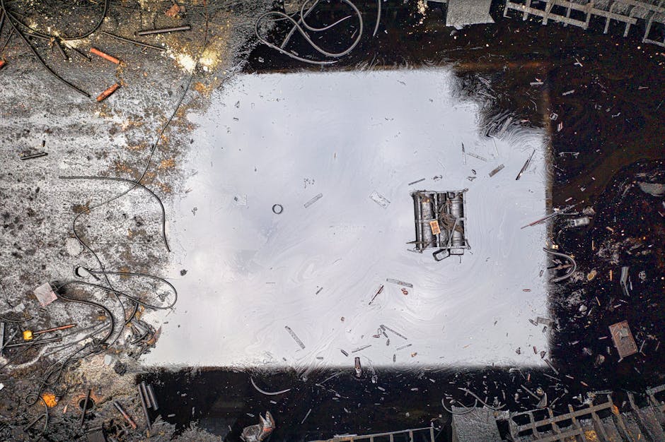 An industrial cleaning scene showing a large, square, stainless steel or metal surface, possibly a floor or a containment area, with a reflective, polished finish. The surface is surrounded by scattered debris, small metal pieces, wires, and tools, indicating recent maintenance work. The area appears to be well-lit with natural or overhead lighting, highlighting the cleanliness and shine of the metal surface. The environment suggests a setting where surface cleaning and hazard management are important, in line with safety standards for hazardous waste. The cleaning service, Cleaner Putney, is associated with maintaining hygiene and safety in such industrial or commercial environments as part of their cleaning and hazard waste compliance offerings.