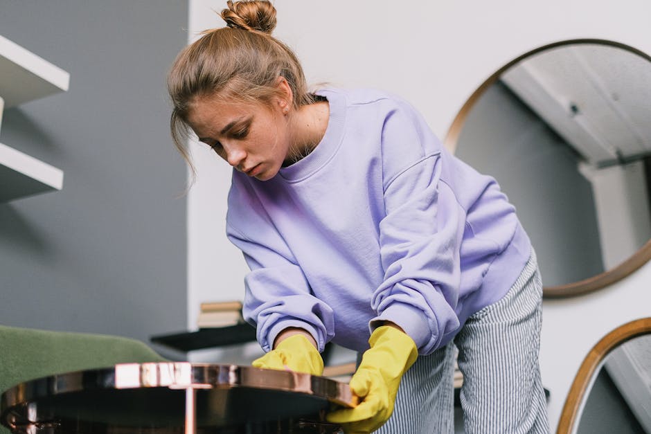 A woman with light brown hair tied in a bun, wearing a lavender sweatshirt and yellow cleaning gloves, is performing surface cleaning on a wooden table in a modern indoor setting. The table has a polished, reflective surface, and she appears focused on wiping or polishing it. The background features a white wall with a round mirror and shelves, as well as a green upholstered piece of furniture, indicating a domestic or office environment. The lighting is bright and even, highlighting the cleanliness of the surface. This image showcases routine cleaning and maintenance typical of professional domestic cleaning services, such as those offered by Cleaner Putney for residential spaces like Putney Pier Apartments.