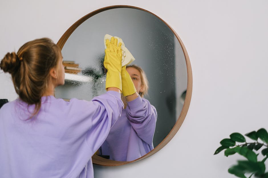 A woman wearing yellow rubber gloves and a lavender long-sleeve shirt is performing surface cleaning on a circular mirror in a residential bathroom. She is using a white cloth to wipe the mirror, removing streaks and water spots, resulting in a clear, streak-free shine. The mirror, framed with wood, reflects her focused expression and the surrounding white wall. The bathroom features a minimalistic design with soft lighting, and a green potted plant is visible in the bottom right corner, adding a touch of natural decor. This image illustrates professional domestic cleaning services by Cleaner Putney, emphasizing detailed mirror and surface sanitisation to maintain hygiene and cleanliness in Putney Pier Apartments, PUTNEY.