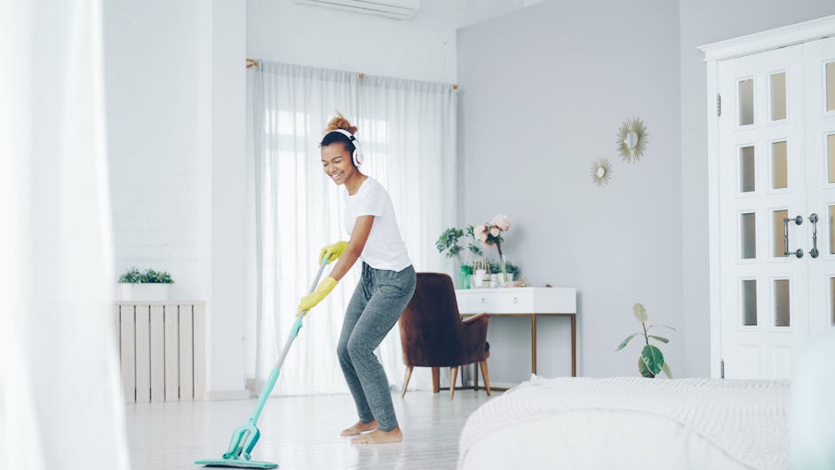 A woman with light brown hair tied in a bun, wearing a lavender sweatshirt and yellow cleaning gloves, is performing surface cleaning on a wooden table in a modern indoor setting. The table has a polished, reflective surface, and she appears focused on wiping or polishing it. The background features a white wall with a round mirror and shelves, as well as a green upholstered piece of furniture, indicating a domestic or office environment. The lighting is bright and even, highlighting the cleanliness of the surface. This image showcases routine cleaning and maintenance typical of professional domestic cleaning services, such as those offered by Cleaner Putney for residential spaces like Putney Pier Apartments.